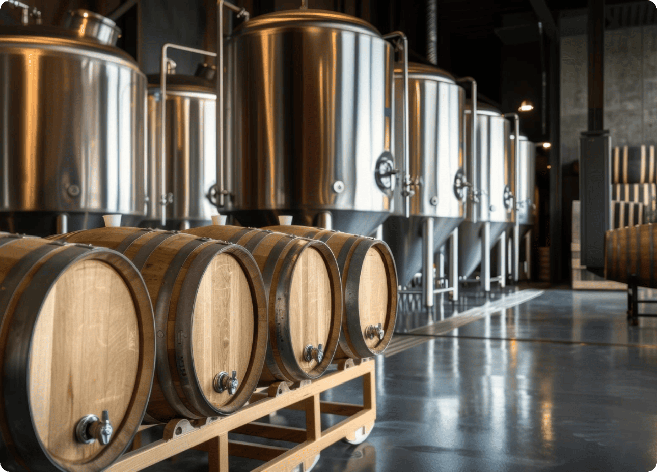 Intérieur d'une brasserie ou distillerie artisanale avec des fûts de chêne au premier plan et des cuves de fermentation en inox à l'arrière.