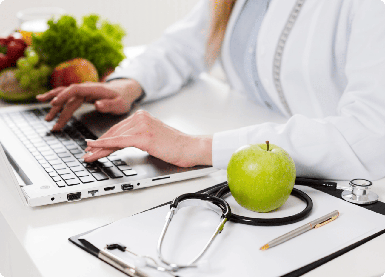 Nutritionniste ou diététicienne en consultation, tapant sur un ordinateur portable à côté d'un stéthoscope, d'une pomme et d'aliments sains.
